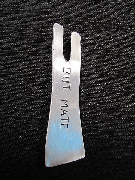 But Mate brooch: Aluminium  Black Singlet shape stamped with the words "BUT MATE"